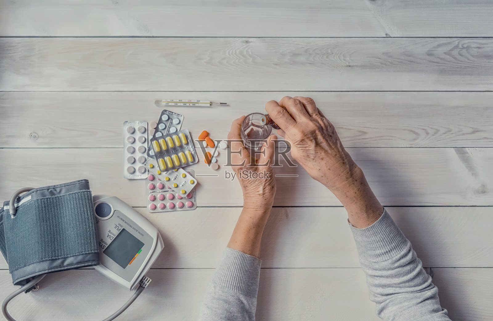 资深手拿药瓶将药水滴入玻璃杯中，药片、眼压计、体温计放在桌上。照片摄影图片