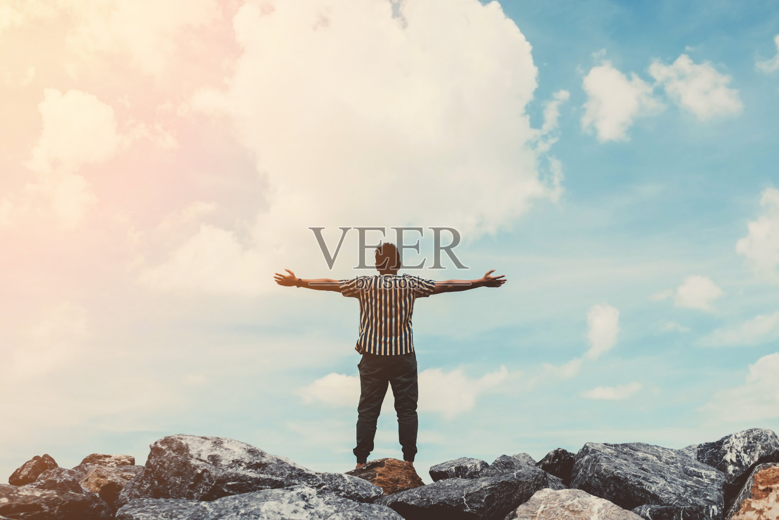 在蓝天白云的抽象背景上，人在岩石顶端举起手。照片摄影图片