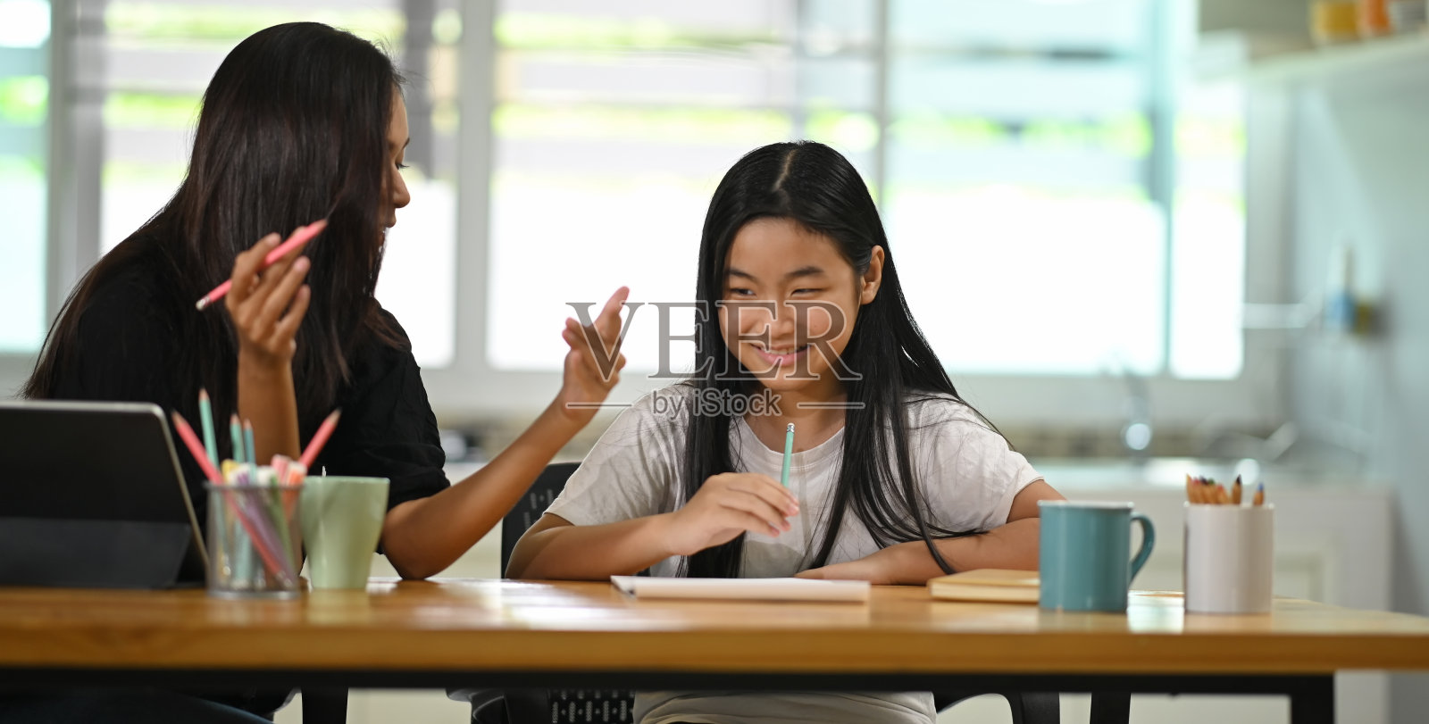 一位年轻的母亲在木桌前教女儿做家庭作业。照片摄影图片