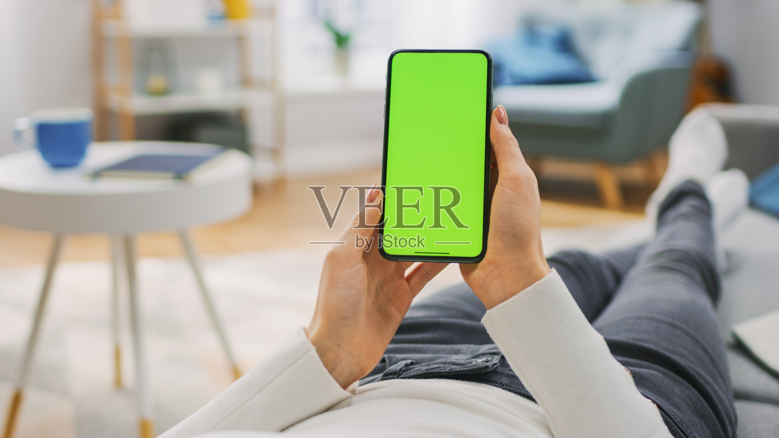 年轻女子躺在沙发上使用绿色模拟屏幕智能手机。女孩使用Chroma Key手机，上网，在社交网络上发帖。视角拍摄。照片摄影图片