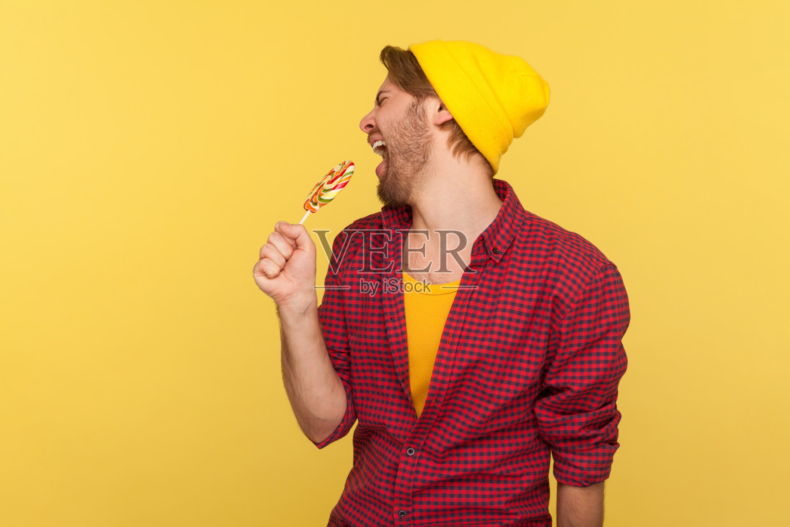 一个无忧无虑的潮人戴着无檐帽，穿着格子衬衫，对着棒棒糖像麦克风一样尖叫。室内照片摄影图片