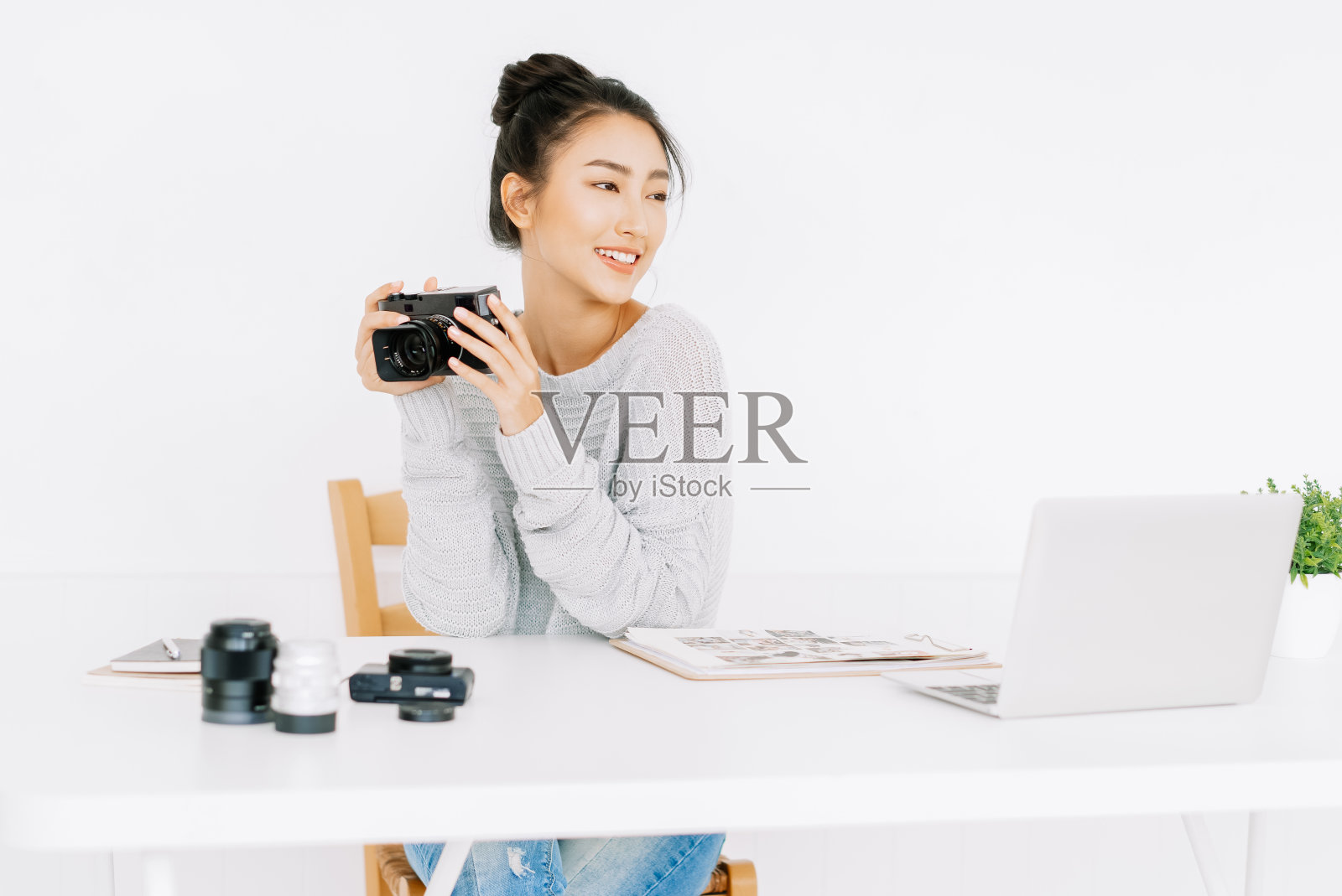 美丽的亚洲女性摄影师的肖像坐在家里办公室的桌子上工作，同时拿着相机，看着一边，脸上带着微笑。自由创业小企业主概念。照片摄影图片
