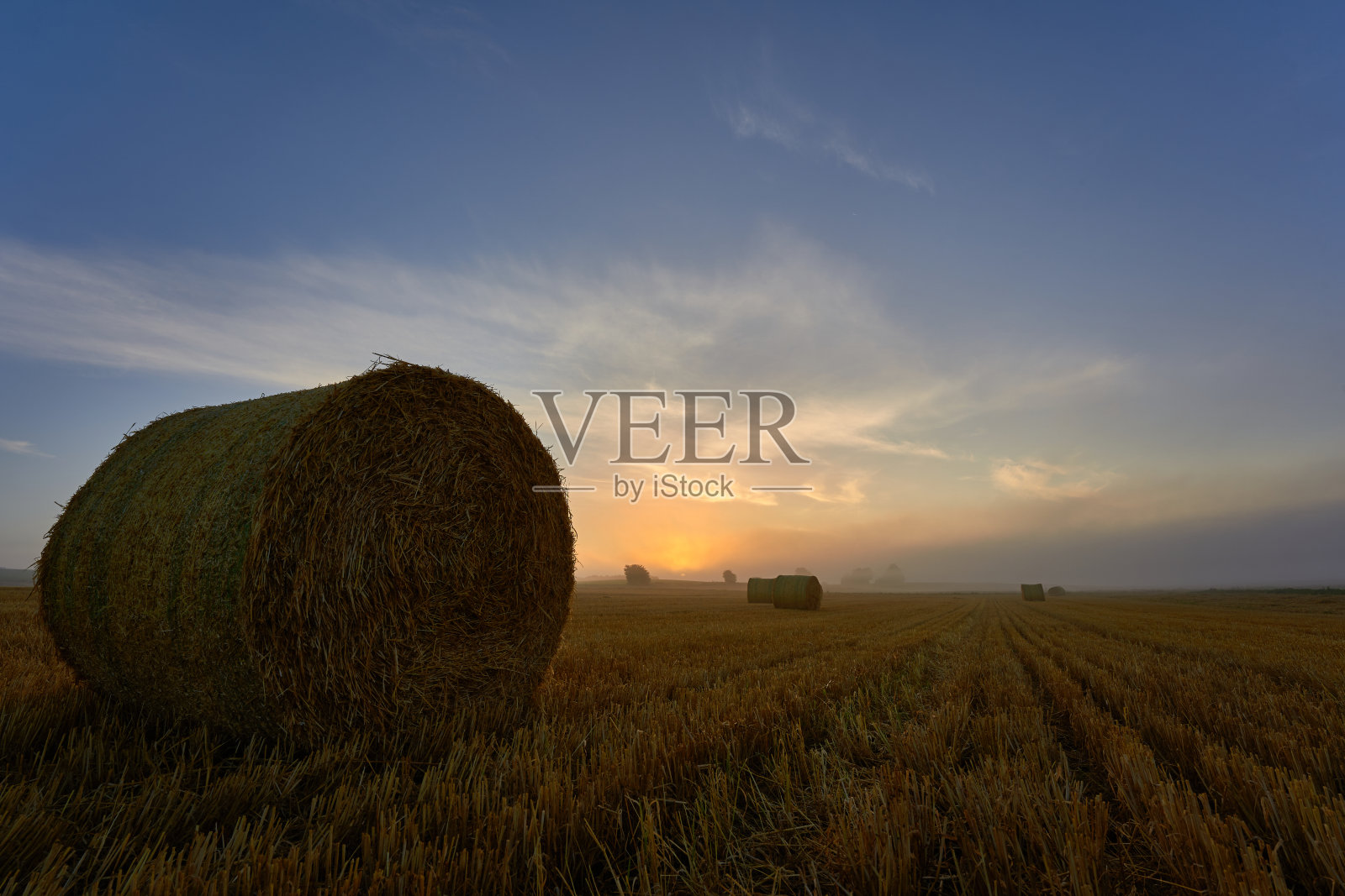 圆圆的干草捆在收获的田野上，明亮的太阳升起，蓝天白云，德国。照片摄影图片
