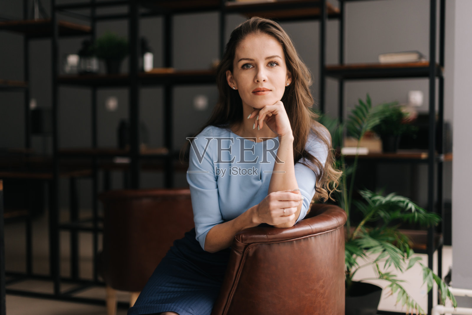 漂亮的年轻女商人穿着时髦的浅蓝色连衣裙坐在办公室的椅子上。照片摄影图片