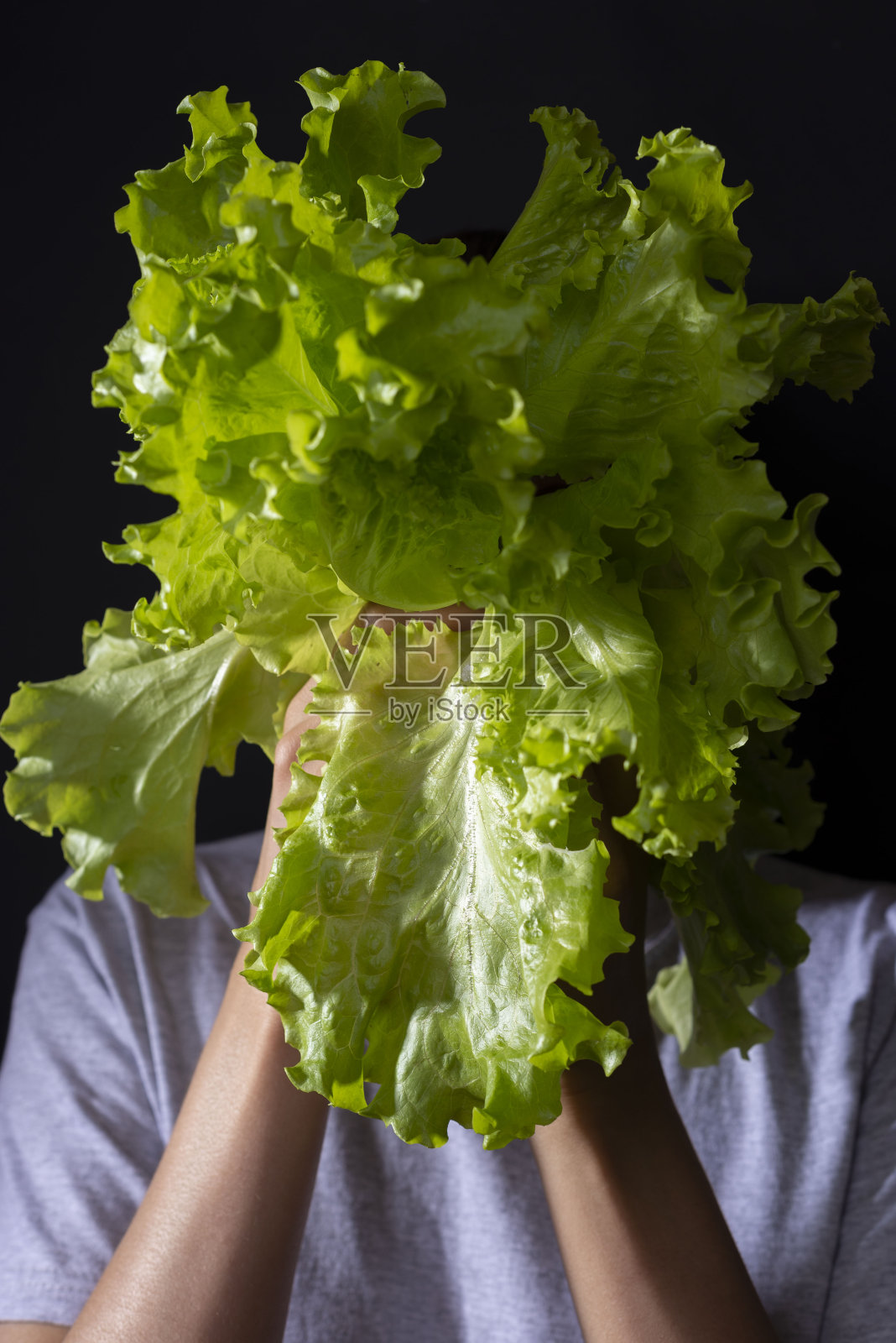 一个女孩用绿色的沙拉叶子遮住脸的肖像。在黑色的背景上。健康食品，蔬菜沙拉中的维生素。新鲜蔬菜。健康营养和素食主义的概念照片摄影图片