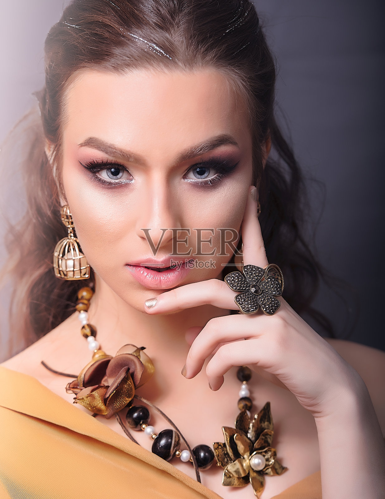 年轻的白人妇女与黄金珠宝的特写。她戴着一条花和石头的声明项链，一对大珍珠耳环和声明花戒指，在灰色的背景照片摄影图片