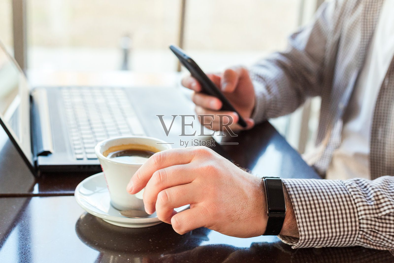 戴着智能手表的男人在工作空间喝咖啡。男人使用笔记本电脑，拿着智能手机进行商务工作或学习。靠近智能手表在男性的手。自由职业者在咖啡馆，办公室，短信按摩照片摄影图片