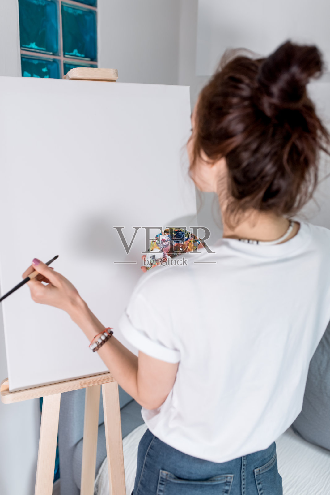 从后面看，她是一名穿着白色t恤的女艺术家，在工作室的家中，手里拿着一支刷子和调色板，上面涂着颜料。背景沙发，画架和白色绘画。照片摄影图片