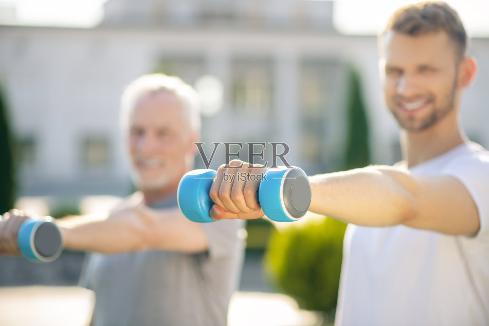 一名留着胡子和灰白头发的年轻男子微笑着在外面练着蓝色哑铃照片摄影图片