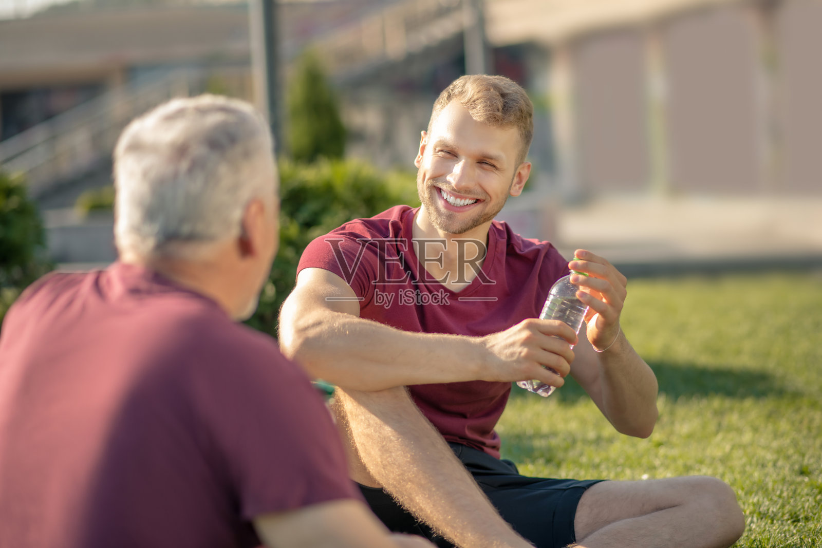 蓄着胡须的男子正合着瓶子，面带微笑，头发灰白的男子坐在他对面的草地上照片摄影图片