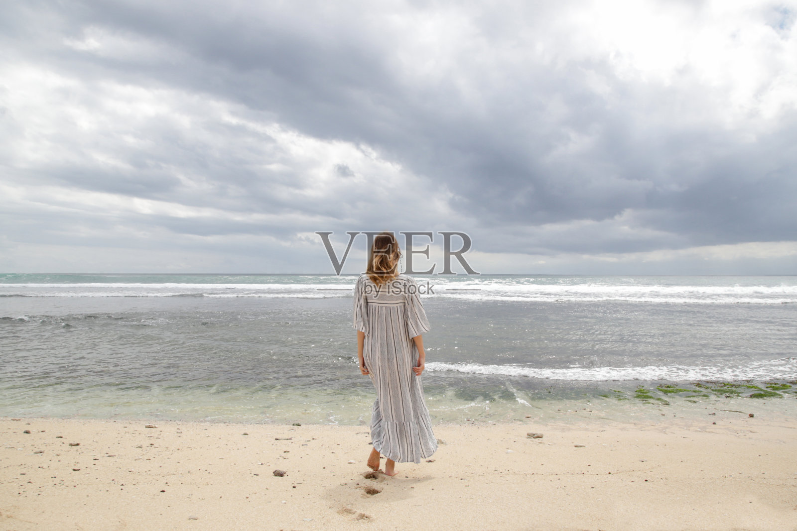 一个女人站在沙滩上，看着地平线。在阴沉的夏日里，一个穿着白色长裙的女人站在岸边。照片摄影图片