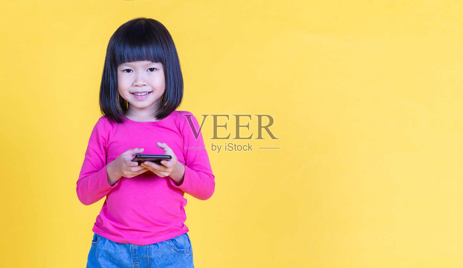 小亚洲女孩的肖像使用智能手机与复制空间隔离在黄色背景。快乐的小学步女孩教育互联网技术，回到学校理念旗帜照片摄影图片