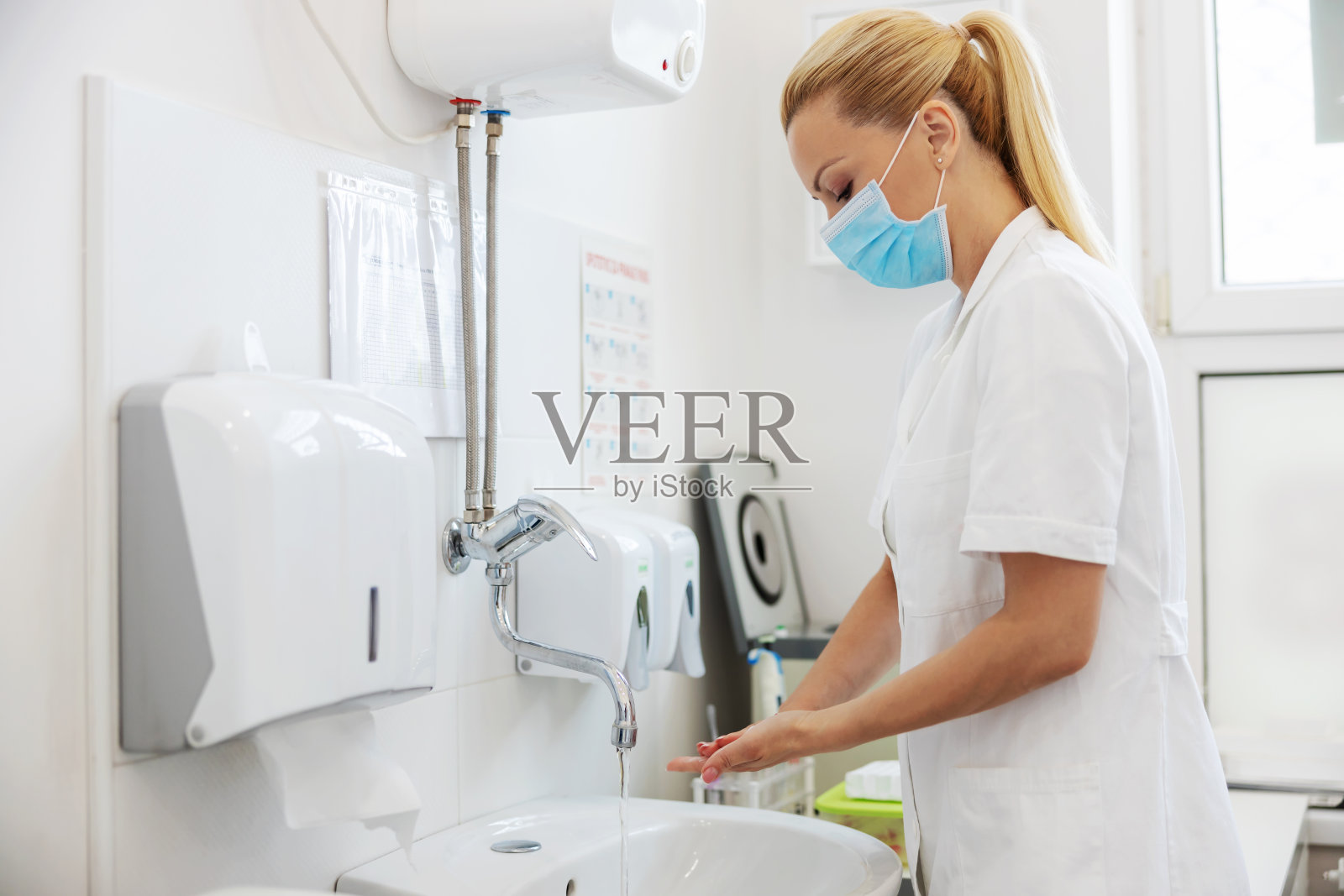 漂亮的金发女实验室助理，穿着无菌制服，脸上戴着防护口罩，在新冠肺炎爆发期间站在休息室洗手。照片摄影图片