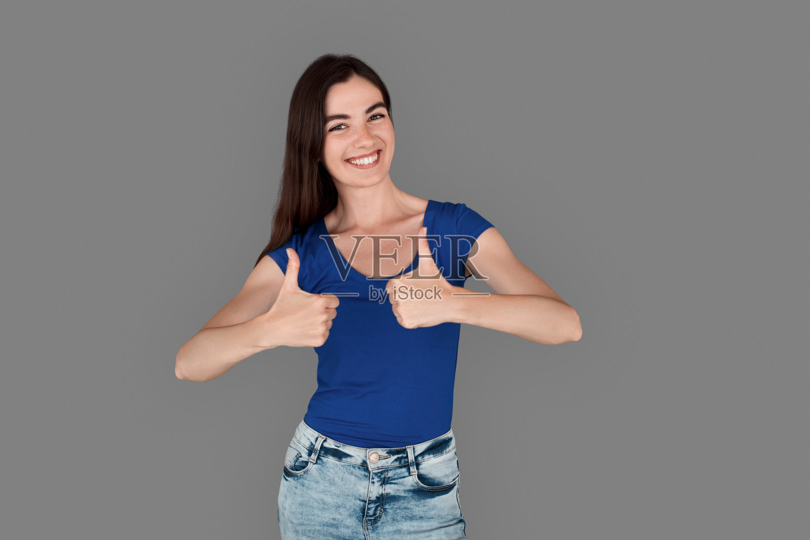 年轻的女孩站在孤立的工作室灰色的大拇指向上笑俏皮照片摄影图片