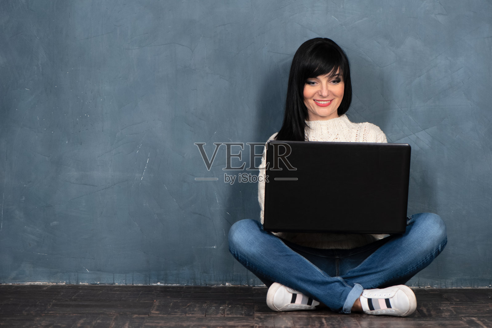 漂亮的黑发女孩坐在地板上，拿着她的笔记本电脑，看着它，微笑着。工作室肖像上的单色蓝色，时髦破旧的背景，文字空间。照片摄影图片