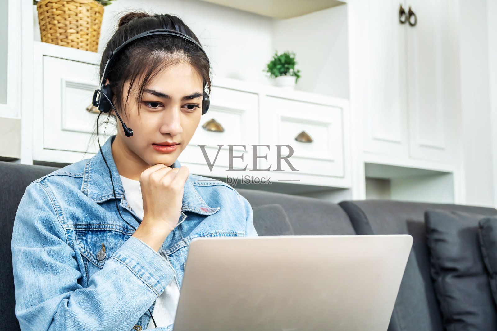 一位年轻的亚洲商业女性正坐在自己家里的沙发上，戴着耳机用笔记本电脑进行视频电话会议，在家里办公，思考新的项目，在家工作照片摄影图片