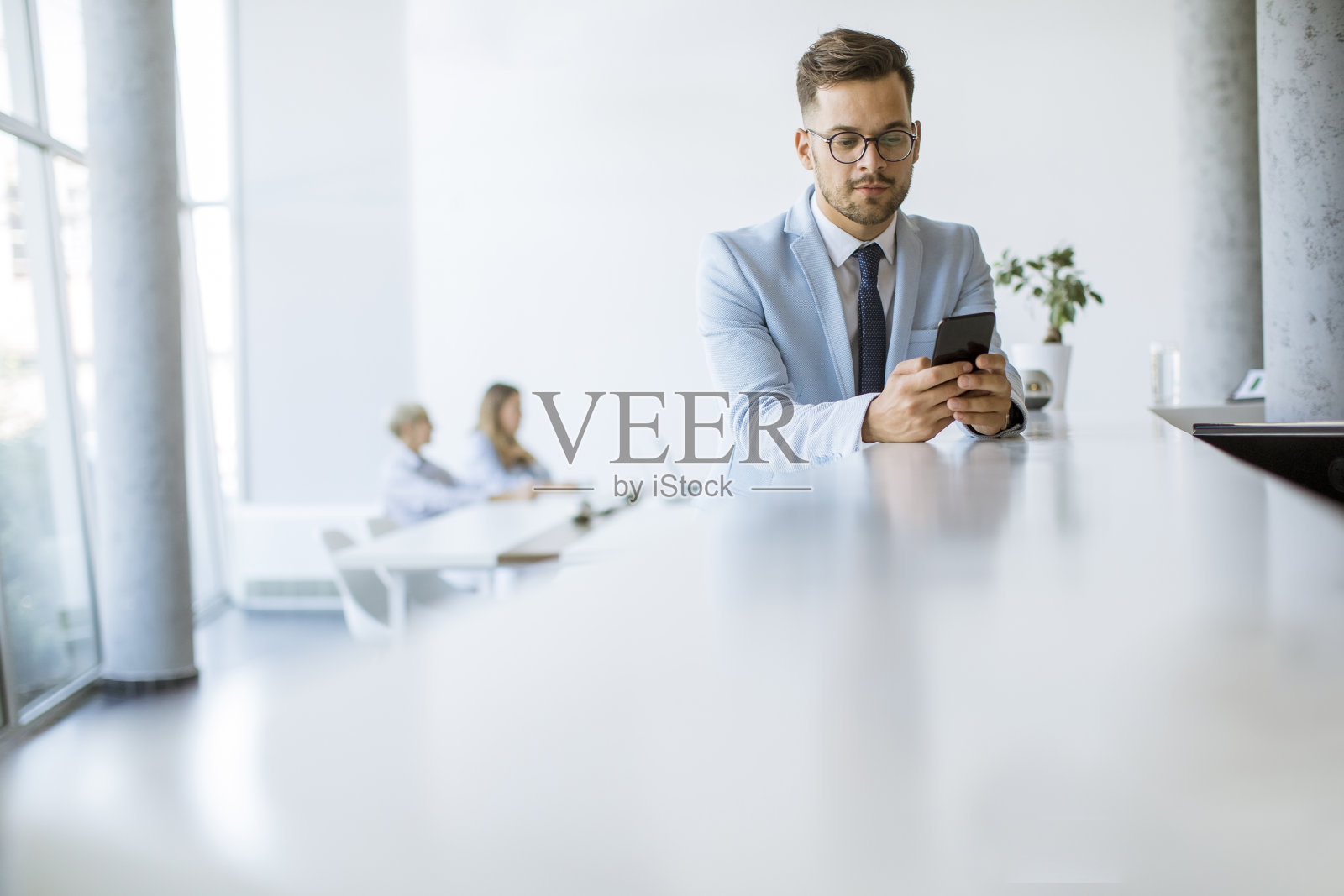 一个快乐的年轻人用他的手机微笑着，而他的同事在后台工作照片摄影图片