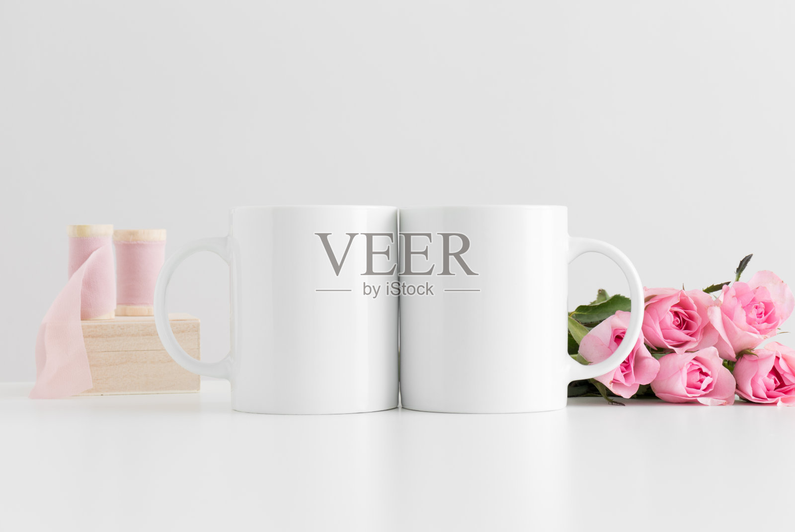 白色的桌子上放着两束粉色玫瑰和丝带的马克杯模型。照片摄影图片