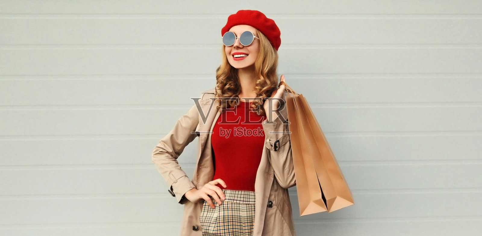 秋天的肖像微笑的女人与购物袋看远穿着大衣，红色的法国贝雷帽在灰色的背景照片摄影图片
