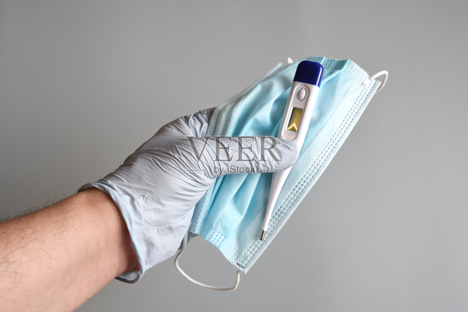 医生双手戴着蓝色手套，手持蓝色外科口罩，灰色背景上佩戴发热温度计，以抵御流行病、疫情、Covid - 19概念。照片摄影图片