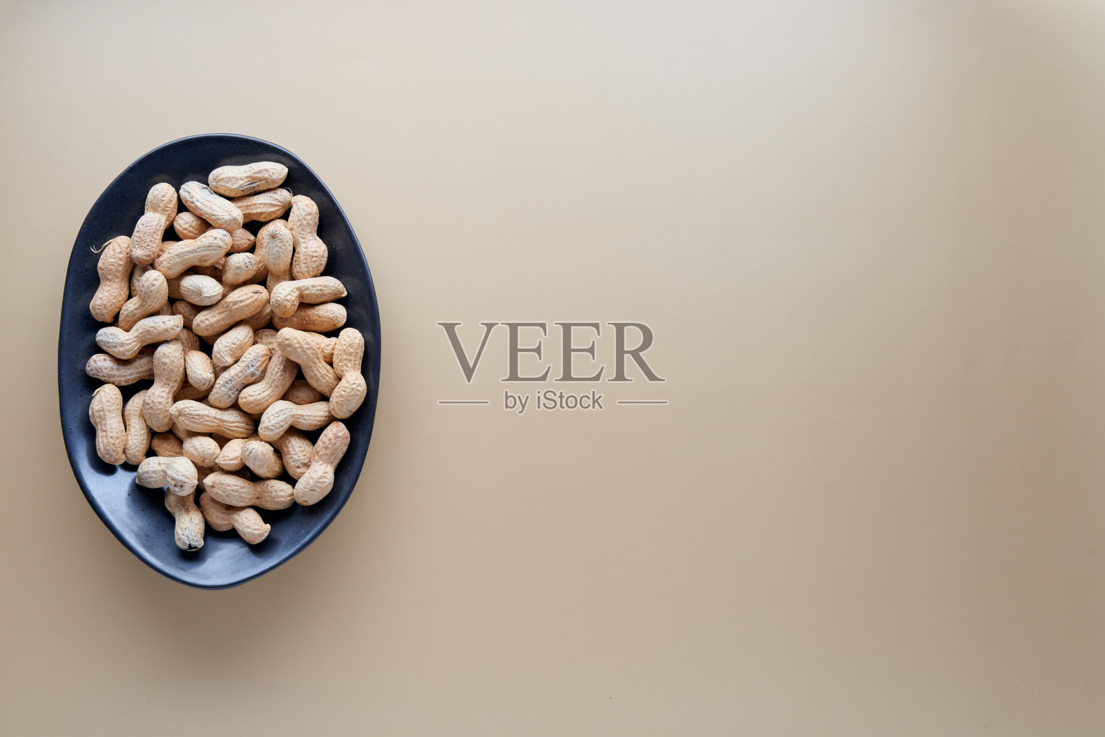 在黑色陶瓷碗中烘烤未剥壳花生，米色背景分离。食品、配料、有机素食蛋白、健康饮食营养理念。顶部视图与复制空间。照片摄影图片