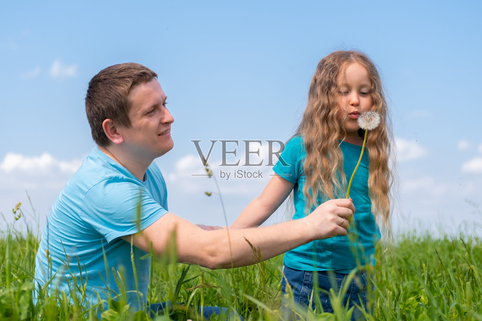 父亲和女儿花时间。夏天，爸爸和女儿们一起在草地上玩耍。女孩吹蒲公英照片摄影图片