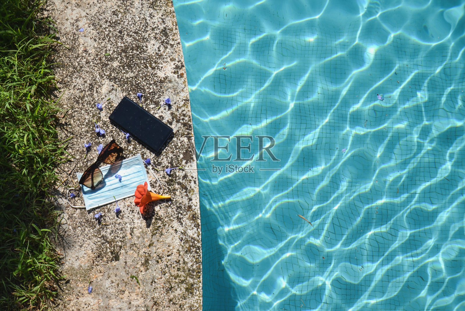 在冠状病毒全球大流行假日期间，放置在游泳池旁的防冠状病毒(COVID-19)的蓝色面罩、太阳镜和手机照片摄影图片