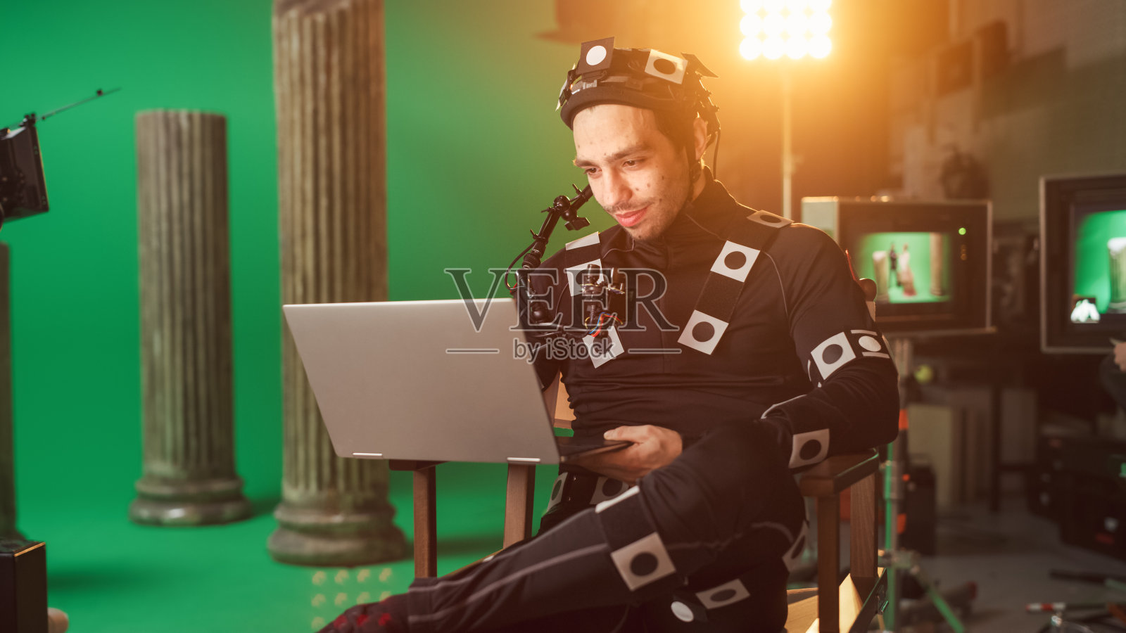 英俊的微笑演员穿着动作捕捉服和头部钻机有午餐休息，坐在椅子上，使用笔记本电脑。高预算电影工作室。关于电影演播室时期的古装剧电影布景照片摄影图片