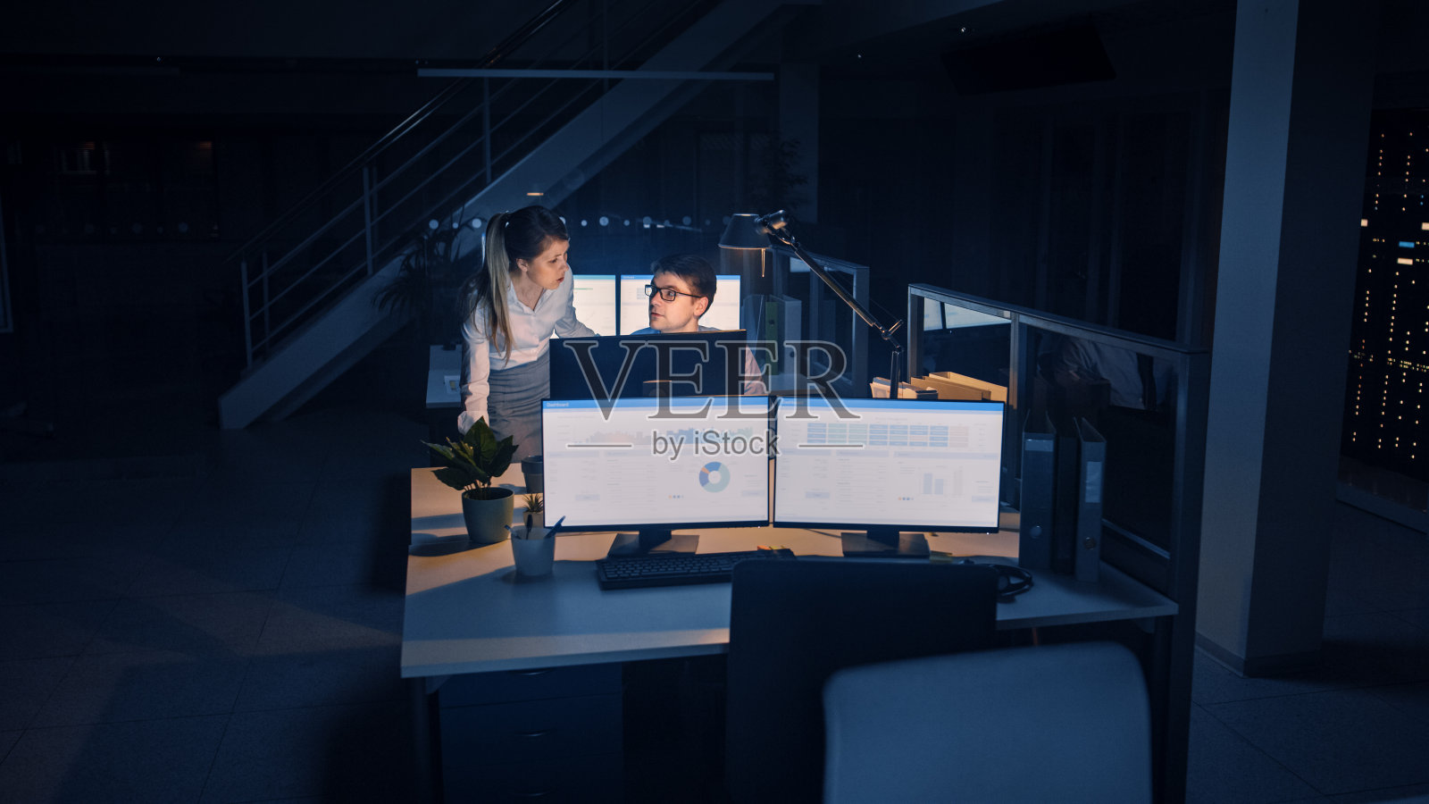 自信的商人使用台式电脑，他的女项目经理解释具体的任务，帐户处理和战略行动。深夜在大公司办公室工作的职场人士照片摄影图片