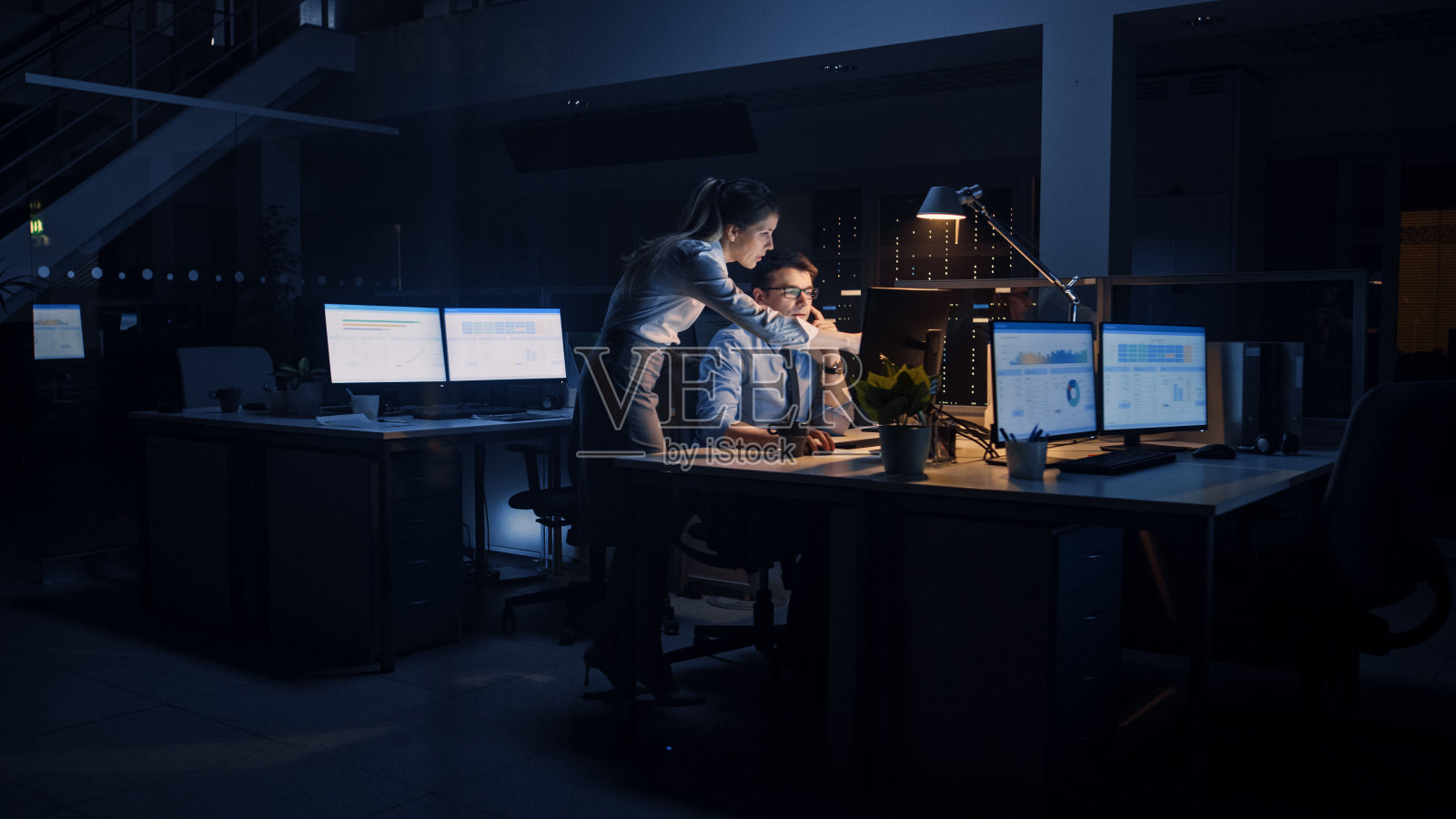 野心勃勃，自信的商人使用台式电脑，他的女性项目经理解释具体的任务，帐户处理和战略行动。深夜在公司办公室工作的职场人士照片摄影图片