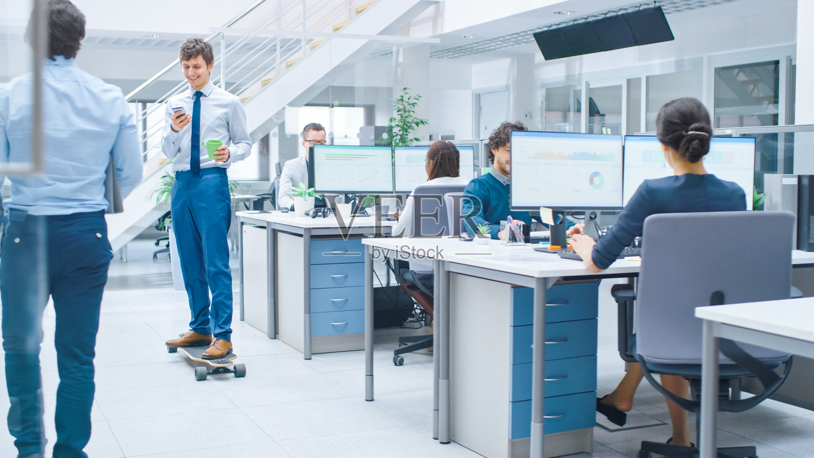 在明亮的公司办公室里，穿衬衫打领带的酷商人在办公室走廊上玩滑板。在后台使用桌面计算机的专业人员照片摄影图片