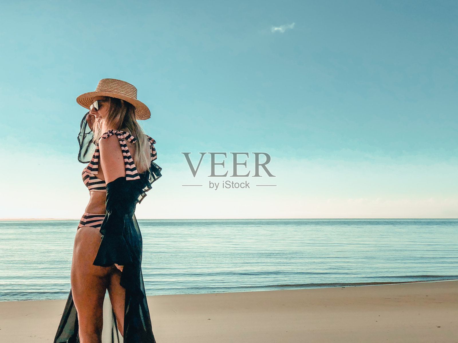 美丽的女人在海滩上在剥离粉红比基尼穿着掩盖与网眼透明泳衣连衣裙和草帽的海洋摄影照片摄影图片