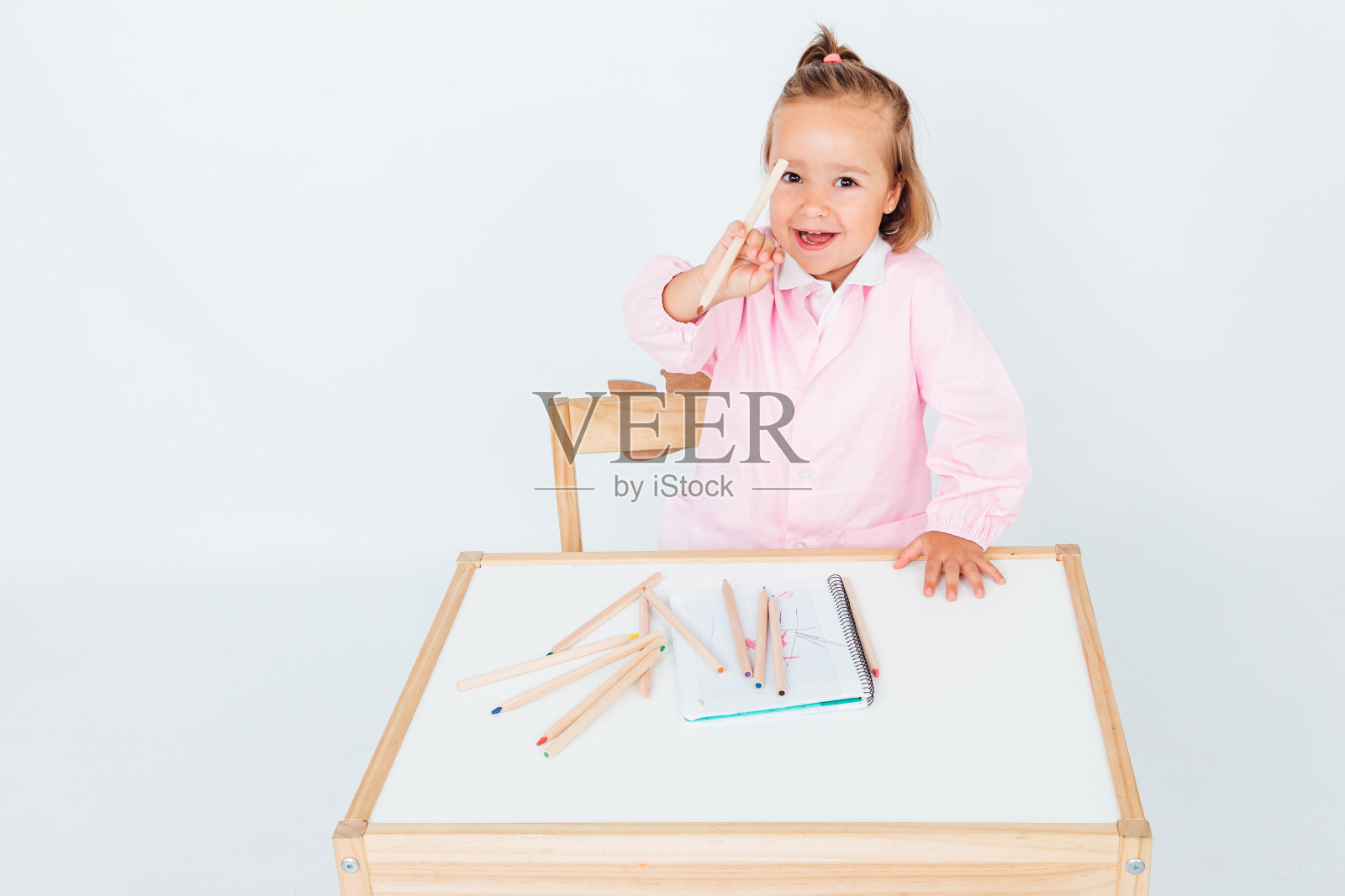 金发女孩，微笑地坐在桌子旁边的椅子上，白色的背景上有笔记本和铅笔。学校的概念照片摄影图片