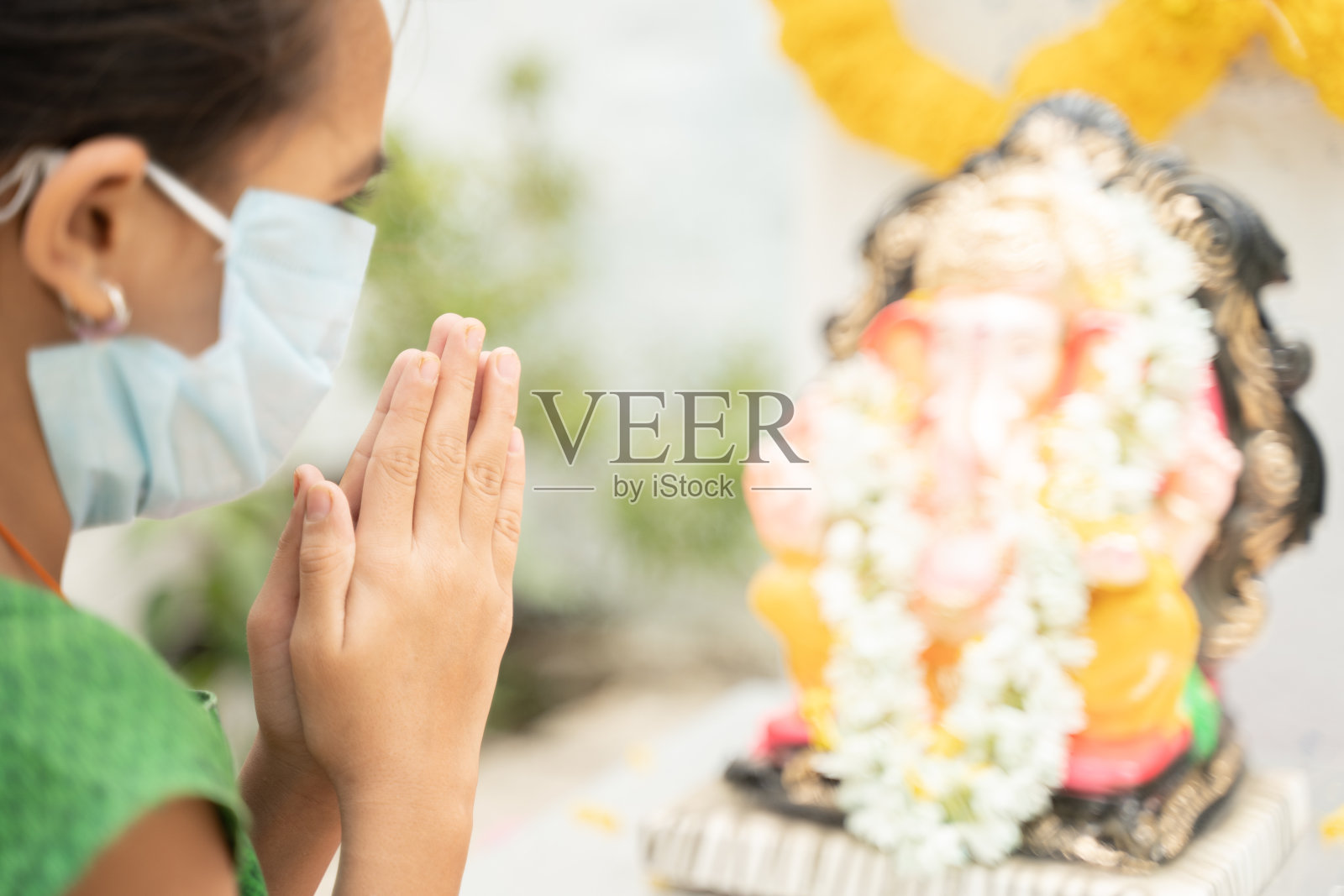 戴医用口罩的女童在象头神节期间闭上眼睛在象头神面前祈祷——covid-19或冠状病毒大流行期间的节日庆祝概念。照片摄影图片