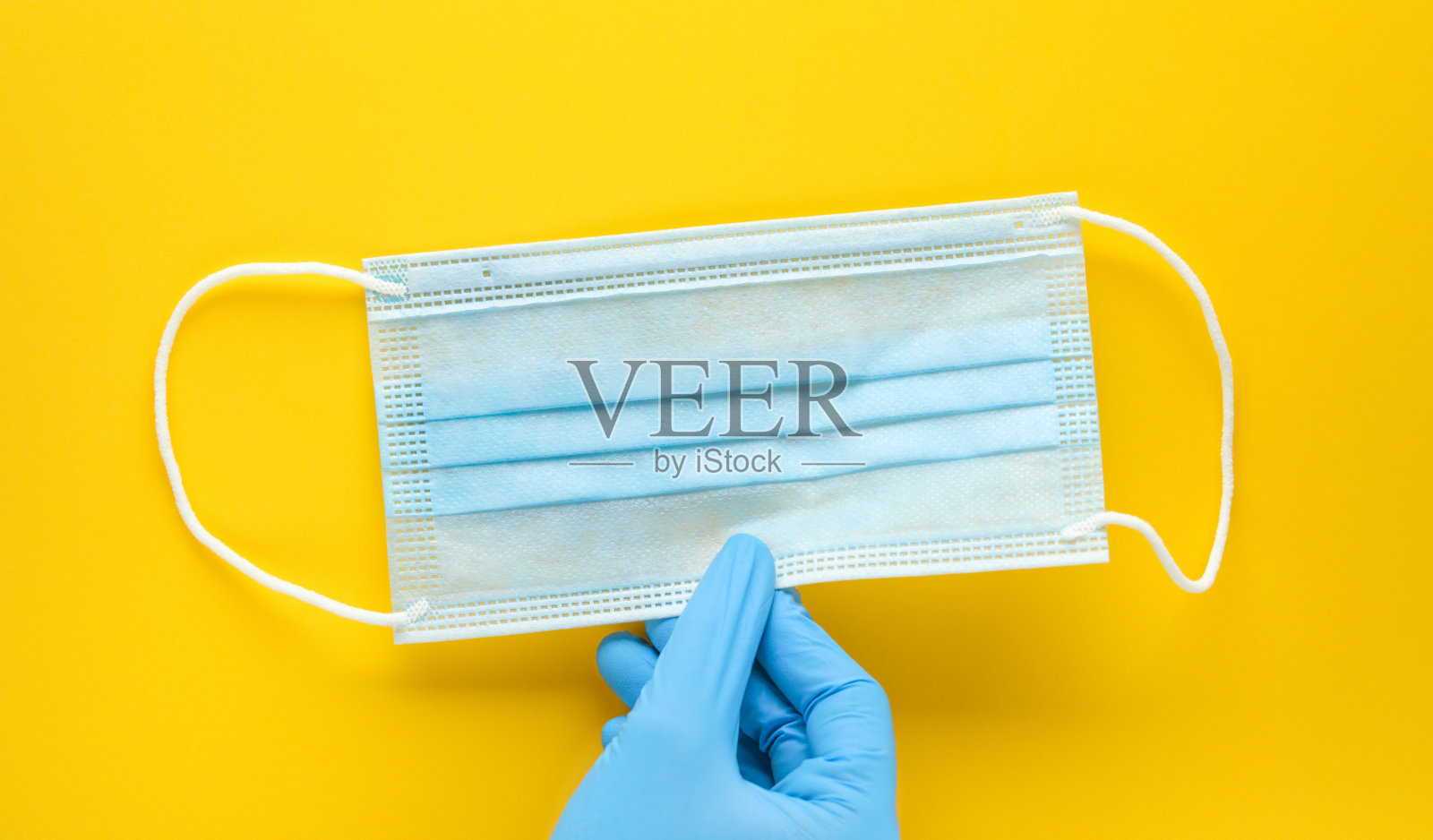 戴着手套的手拿着一个一次性医用口罩，背景为黄色。个人防护装备，PPE。照片摄影图片