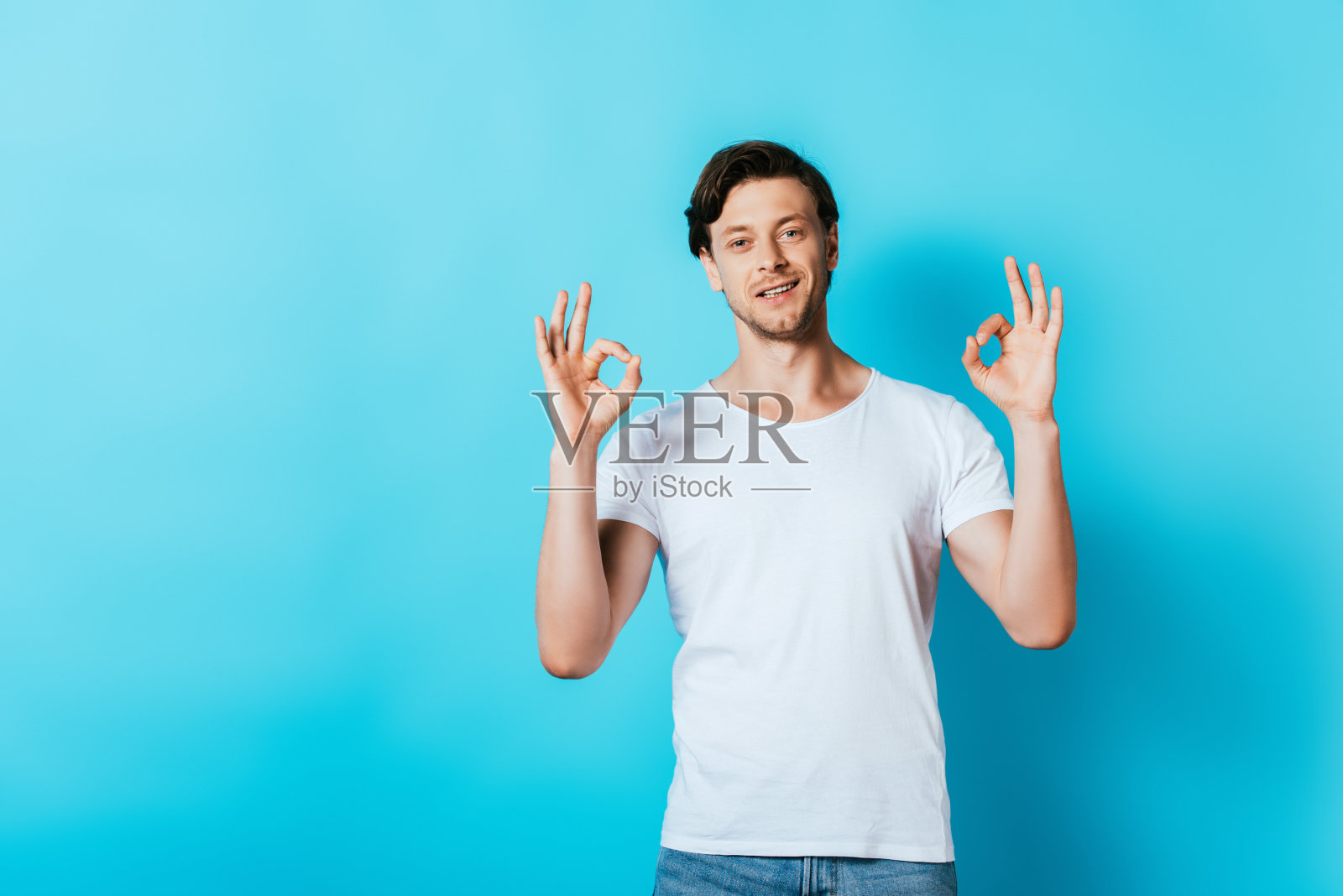 一个穿着白色t恤的年轻人在蓝色背景下对着相机做手势照片摄影图片