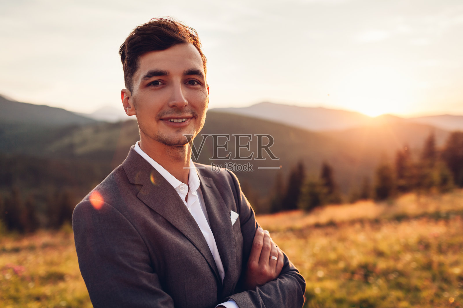 夕阳下，英俊的新郎穿着婚纱走在山间。微笑的男人双手交叉欣赏风景照片摄影图片