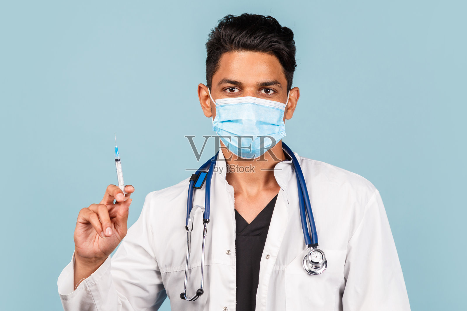 可爱的印度/亚洲医生，手里拿着听诊器和注射器，穿着白色外套，背景是蓝色的。空间的文本照片摄影图片