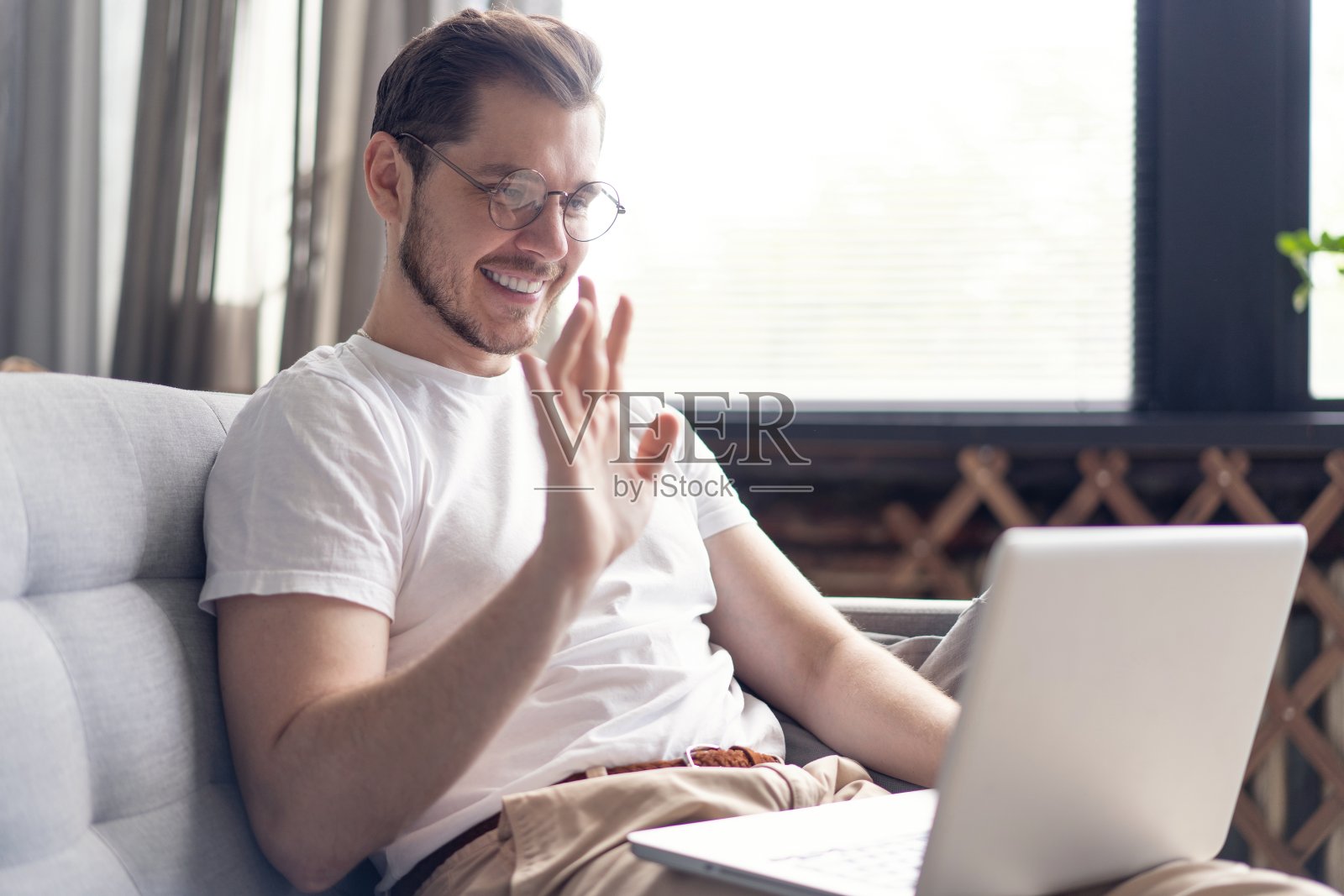 一个快乐的年轻人坐在家里的沙发上用笔记本电脑视频聊天。照片摄影图片