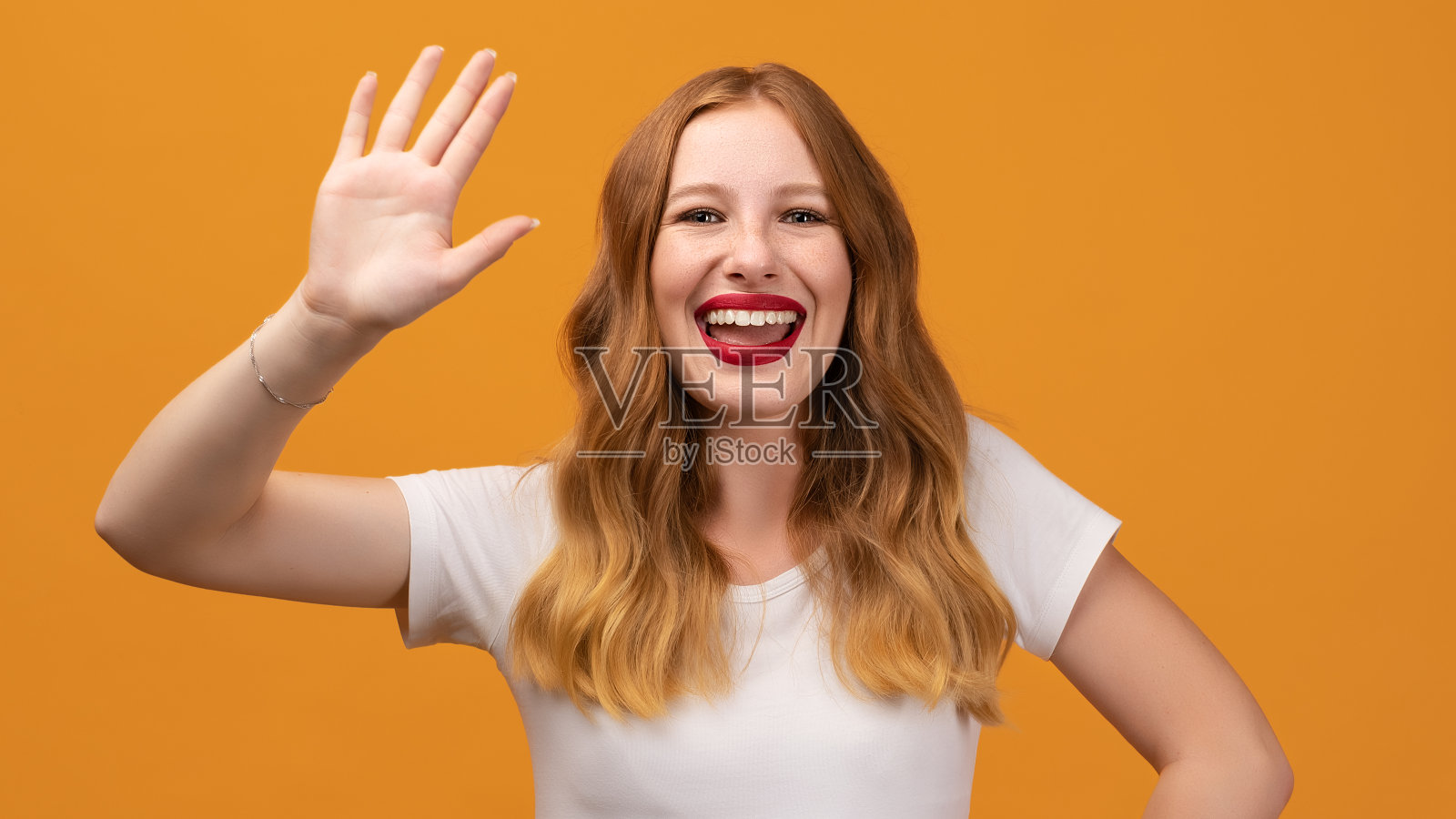 友好的女人的肖像与波浪红头发，站在挥手，看着镜头与迷人的牙齿微笑照片摄影图片