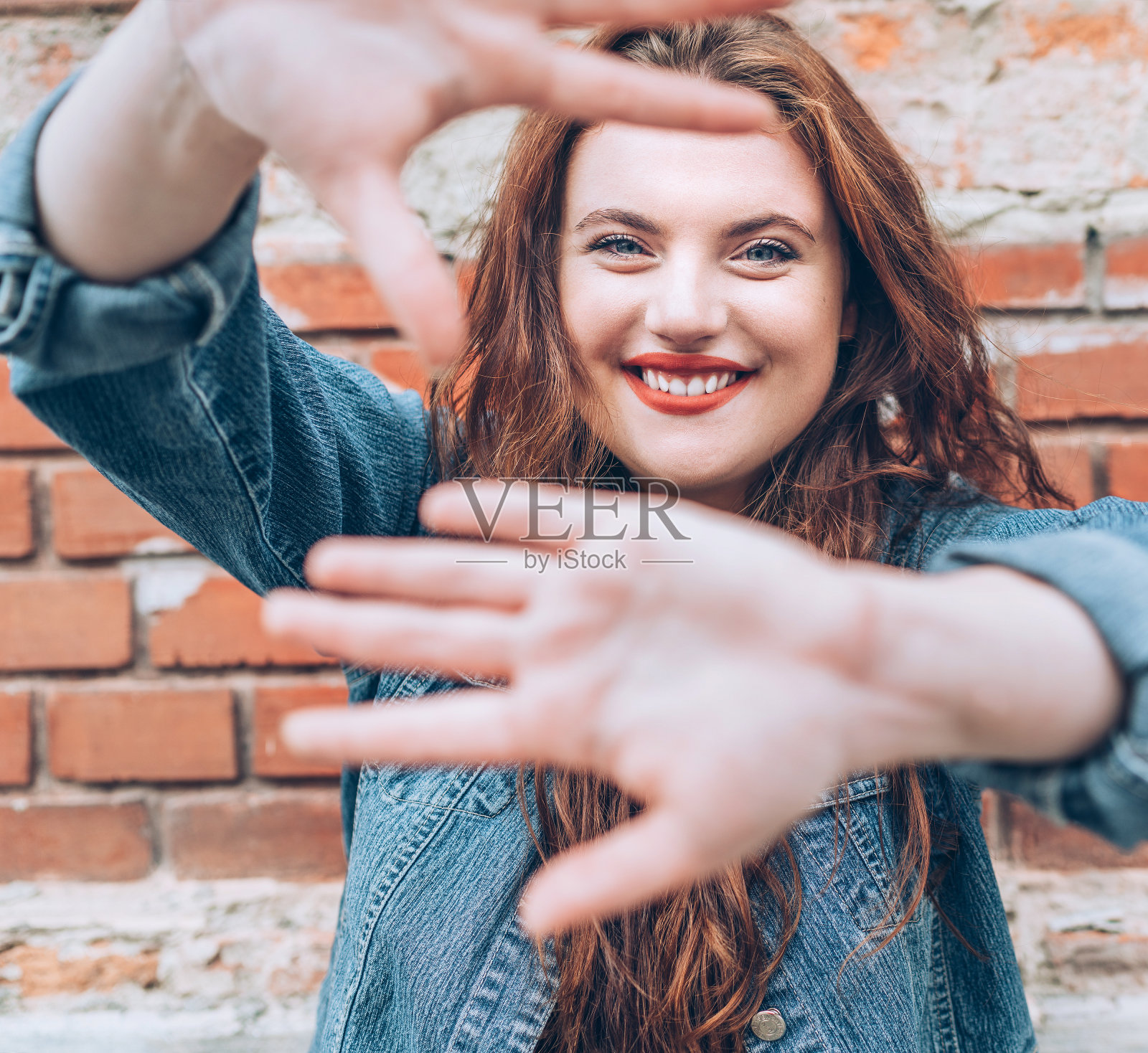 这是一幅真诚微笑的红色鬈发白种人女子的肖像，她涂着红色口红，有着蓝色的眼睛，以红色砖墙为背景，做着“不拍照”的手势。照片摄影图片
