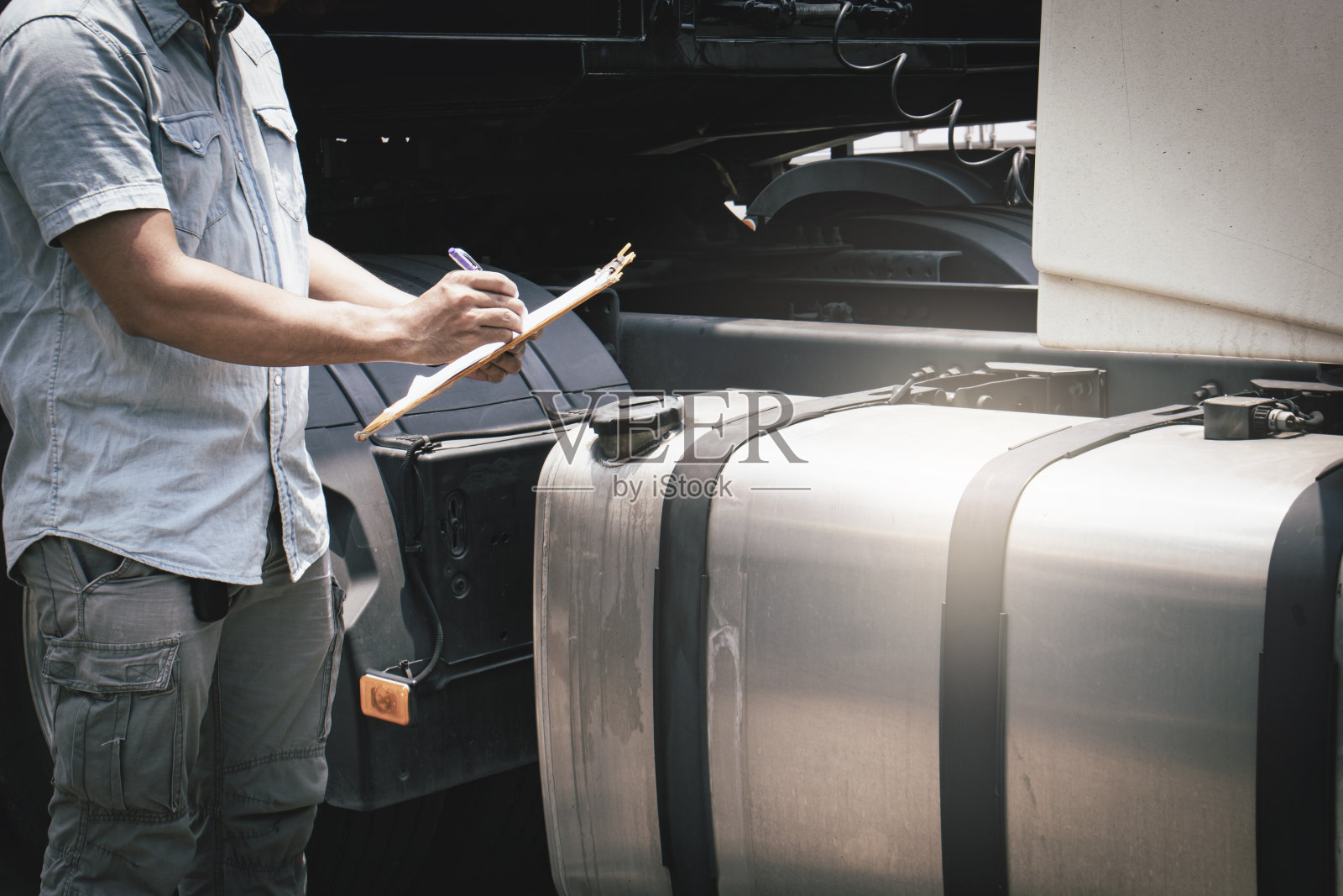 一名手持剪贴板的卡车司机正在检查半挂车的大油箱的安全。照片摄影图片