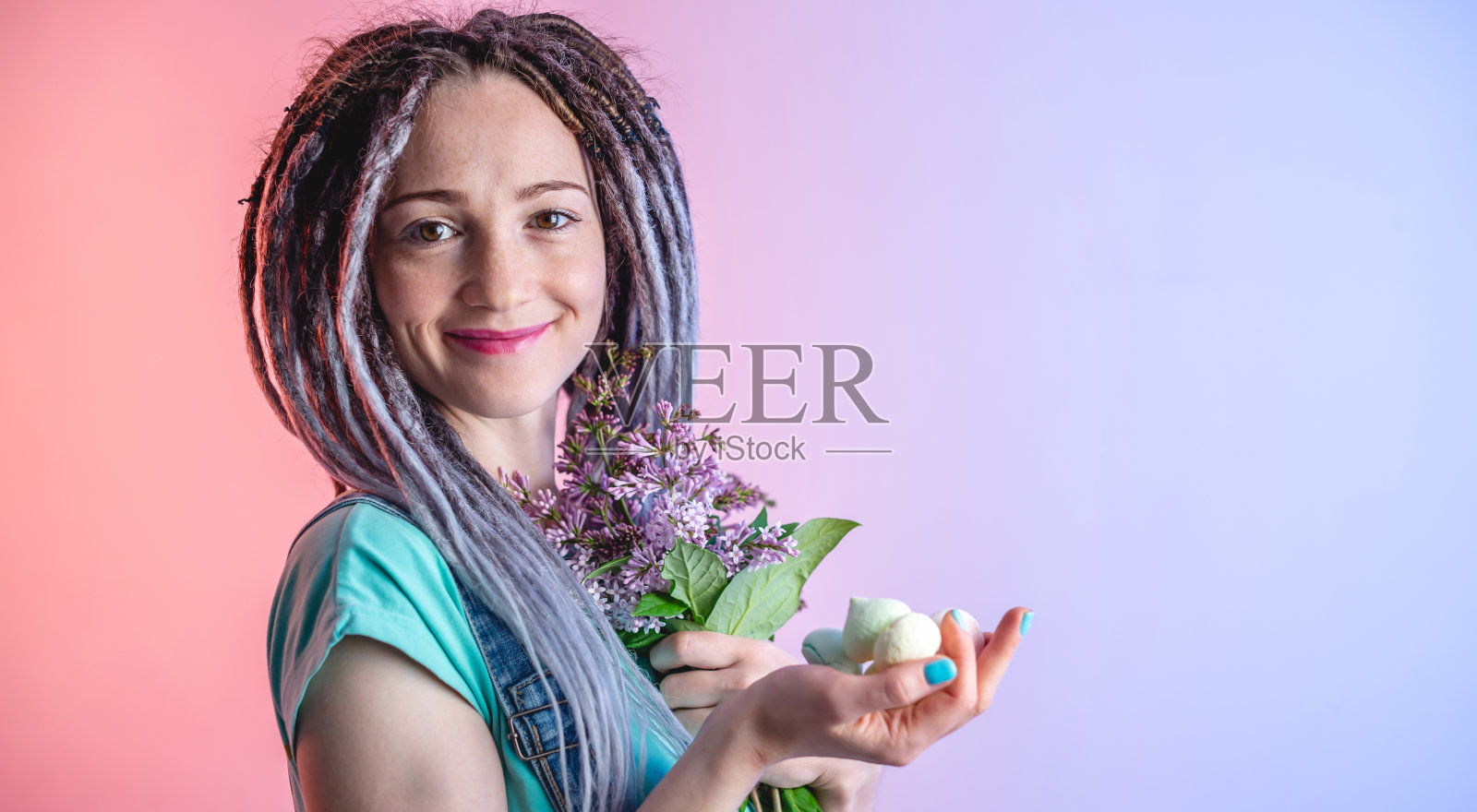 一个梳着发辫的年轻快乐的女人，手里拿着一束美丽的紫丁香花，背景是蓝色和粉红色。一种欢快的心情的概念照片摄影图片
