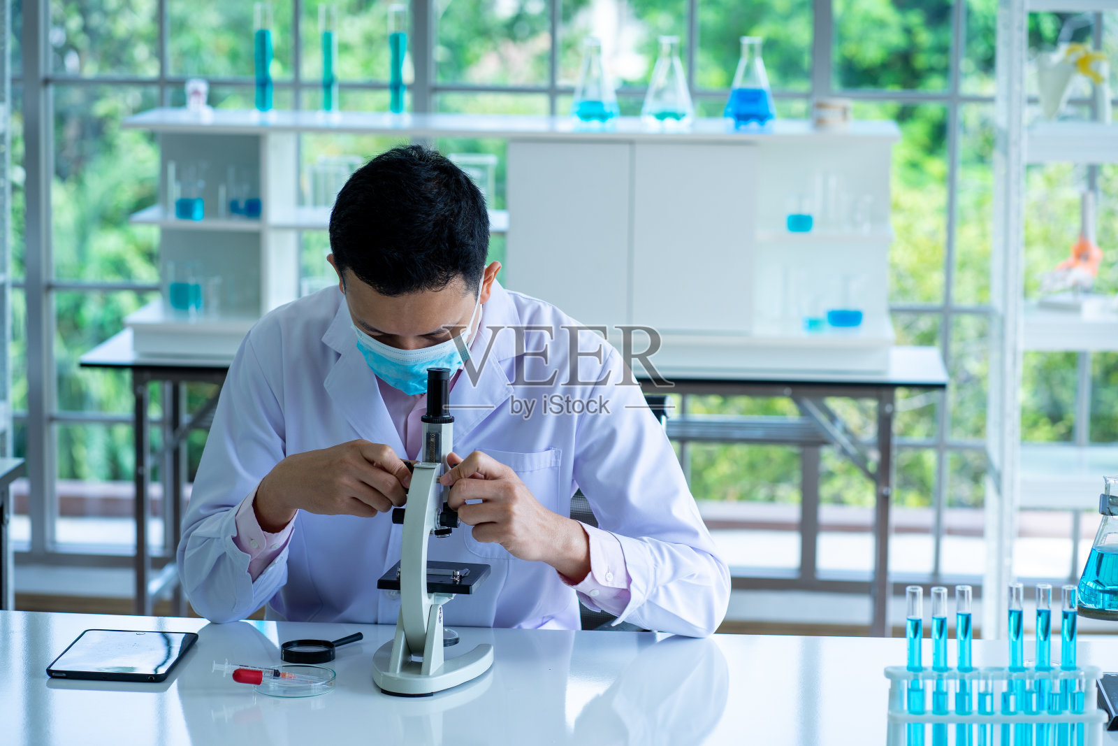 在临床实验室使用显微镜时戴防护外科口罩和实验服的医学科学家照片摄影图片