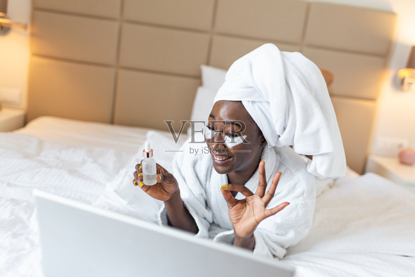 美丽的年轻非洲妇女在浴衣显示她的油滴血清在视频通话与她的朋友在她的笔记本电脑。看着笔记本电脑和她的朋友通过视频通话。照片摄影图片