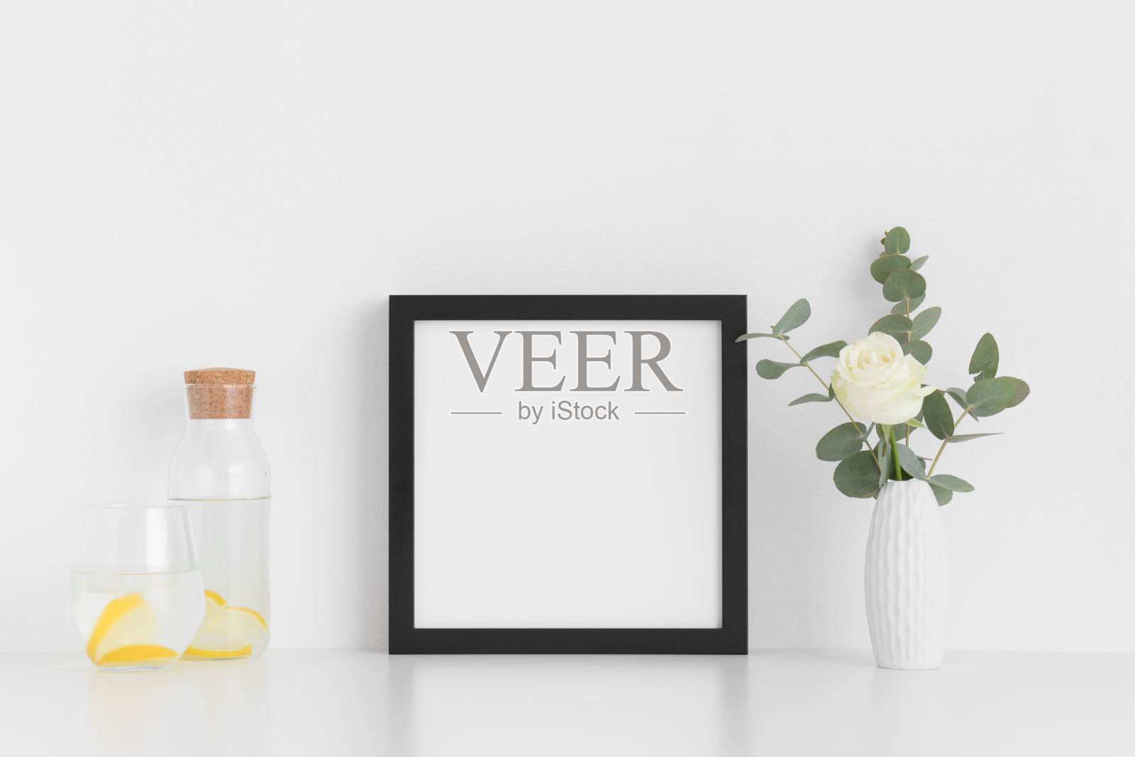 黑色方形框架模型与玫瑰和桉树在花瓶，玻璃和一个瓶子在白色的桌子上。照片摄影图片