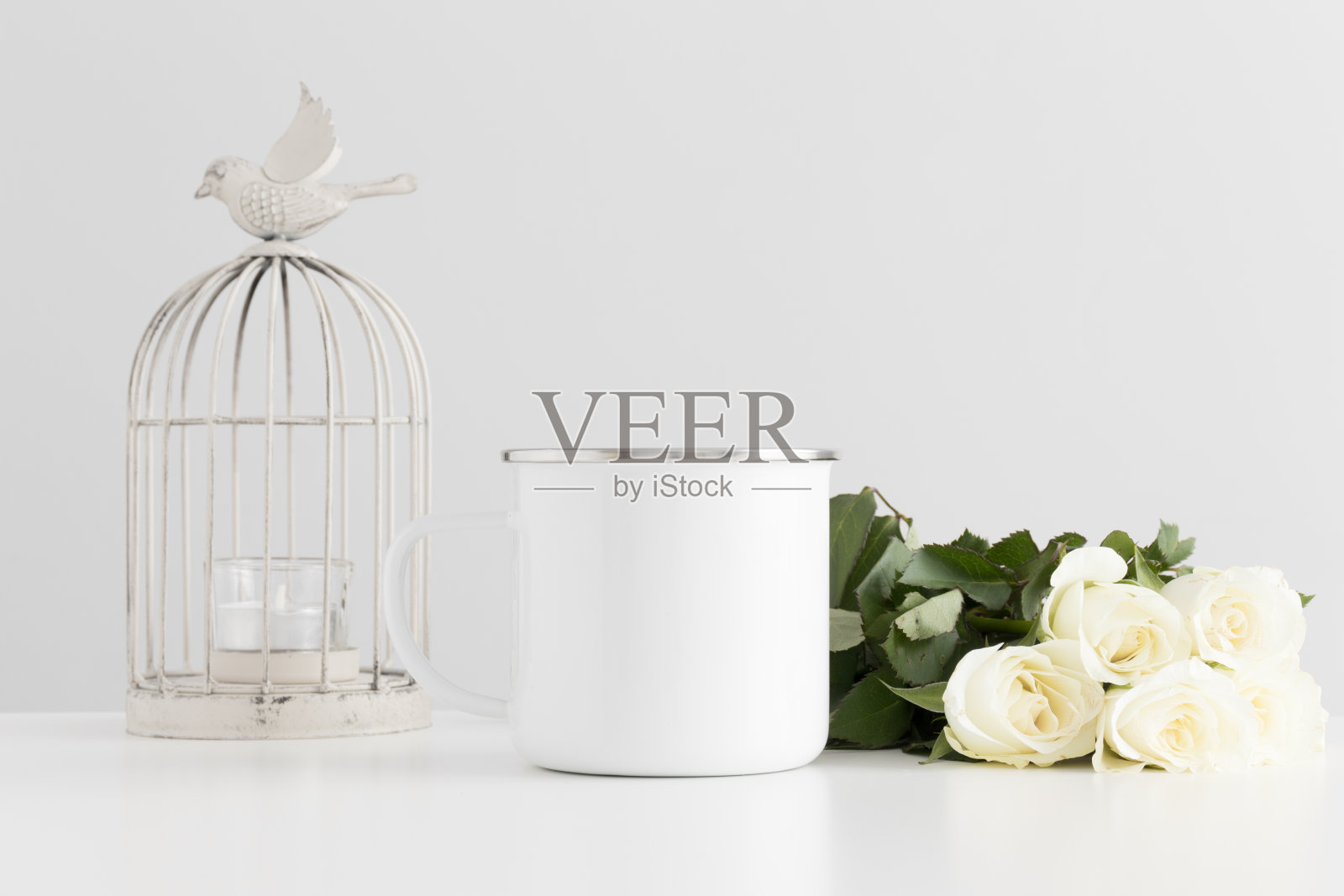 搪瓷杯模型与一束玫瑰和烛台上的白色桌子。照片摄影图片