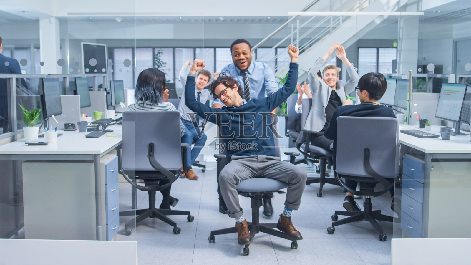 快乐的员工推着他英俊的同事坐在一排排办公桌之间的椅子上，不同的商务人士在现代办公空间的台式电脑上工作。照片摄影图片