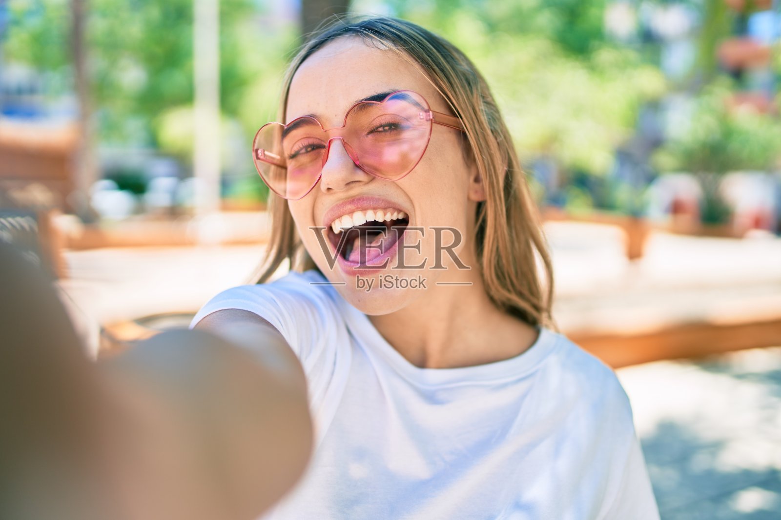 年轻美丽的金发白种女人微笑在一个阳光明媚的日子戴着心形太阳镜在户外自拍照片摄影图片