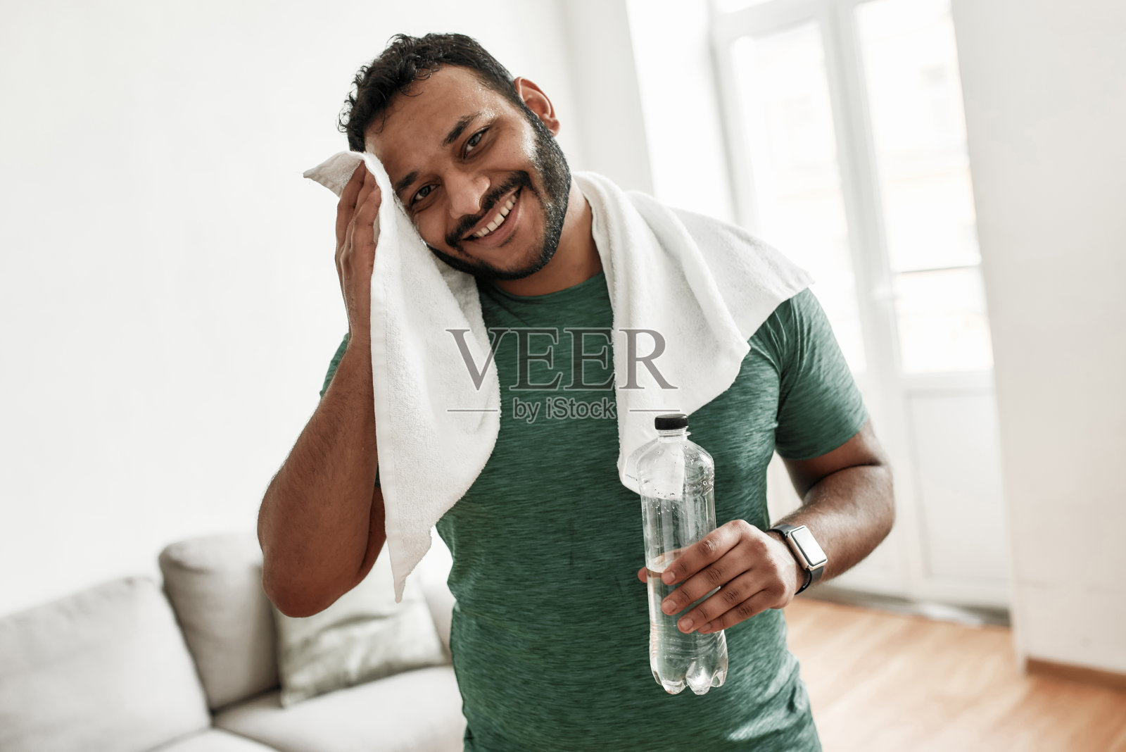 很累,但是很开心。年轻男子在训练锻炼后在家休息时，对着镜头微笑，拿着水瓶，用毛巾擦汗。运动,健康的生活方式照片摄影图片